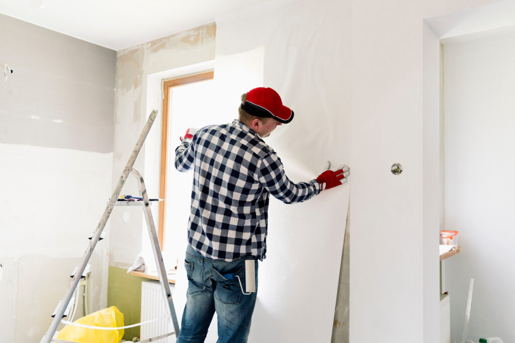 A person wearing a red cap and gloves is standing on the floor by a ladder, applying wallpaper to a wall in a room under renovation. There are renovation tools and supplies visible in the background.