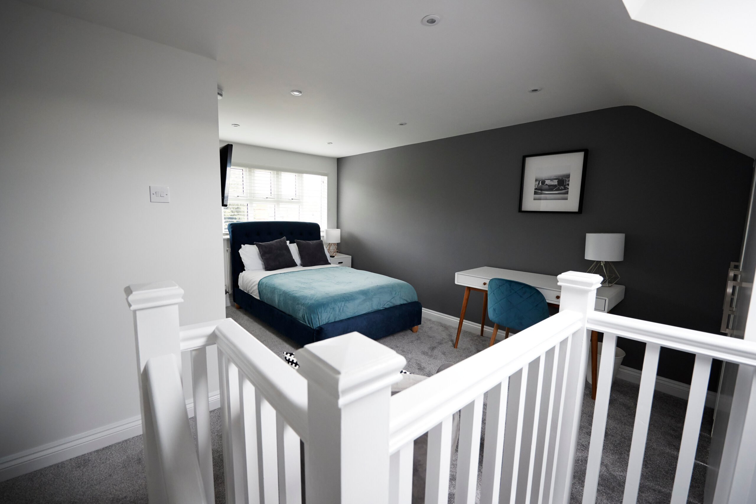 A modern bedroom with gray walls, a double bed with a blue headboard and teal bedding, a white desk with a lamp and blue chair, carpeted floor, and a white railing in the foreground.