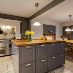 Modern kitchen with gray cabinets, wooden countertops, and stainless steel appliances. A vase of yellow flowers and jars of fruit sit on the island. Dining area with wooden table and chairs in the background. Bright lighting.