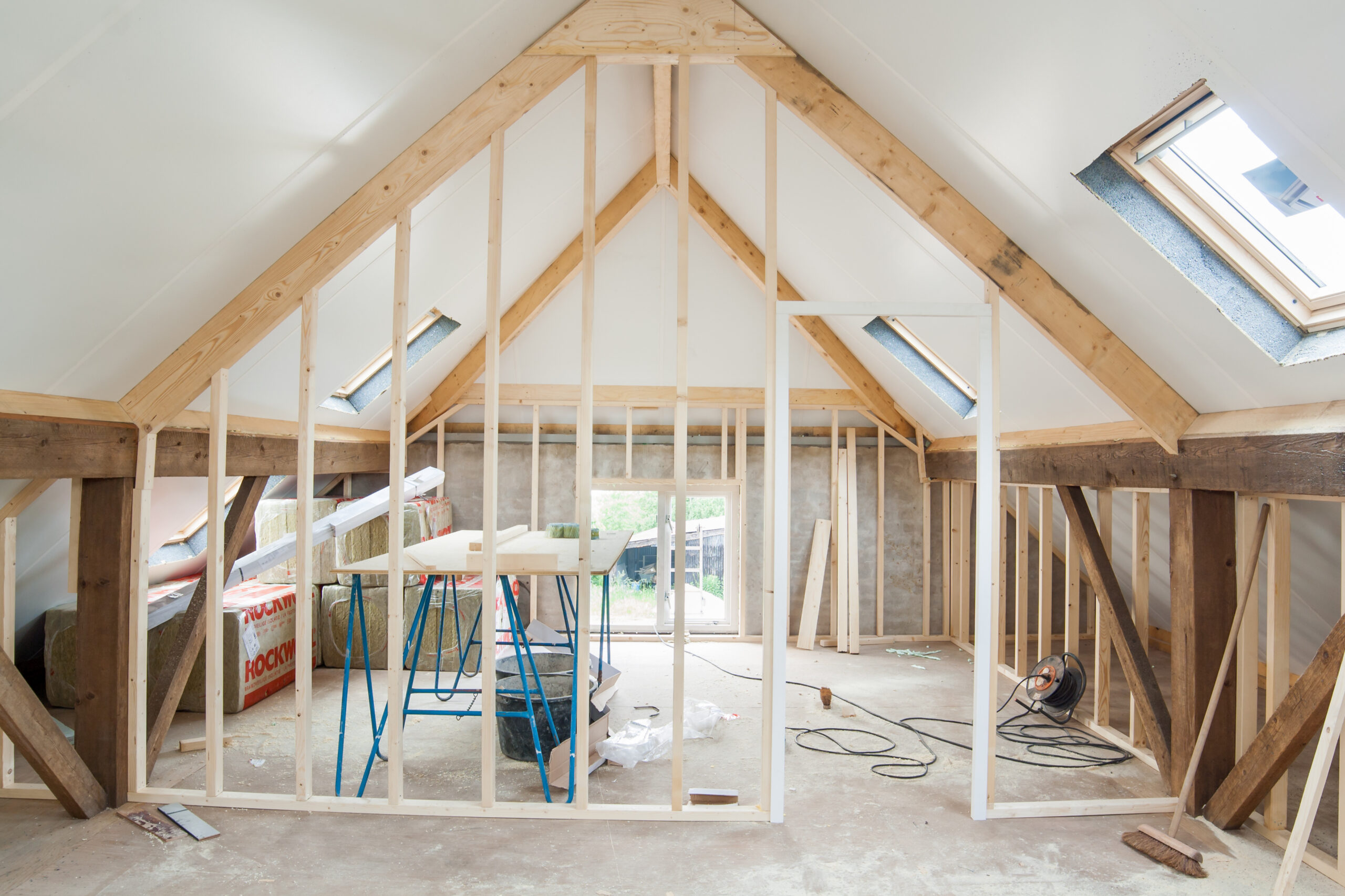 An attic space under renovation with exposed wooden framing, construction materials, tools, and skylight windows on both sides, allowing natural light into the unfinished room.
